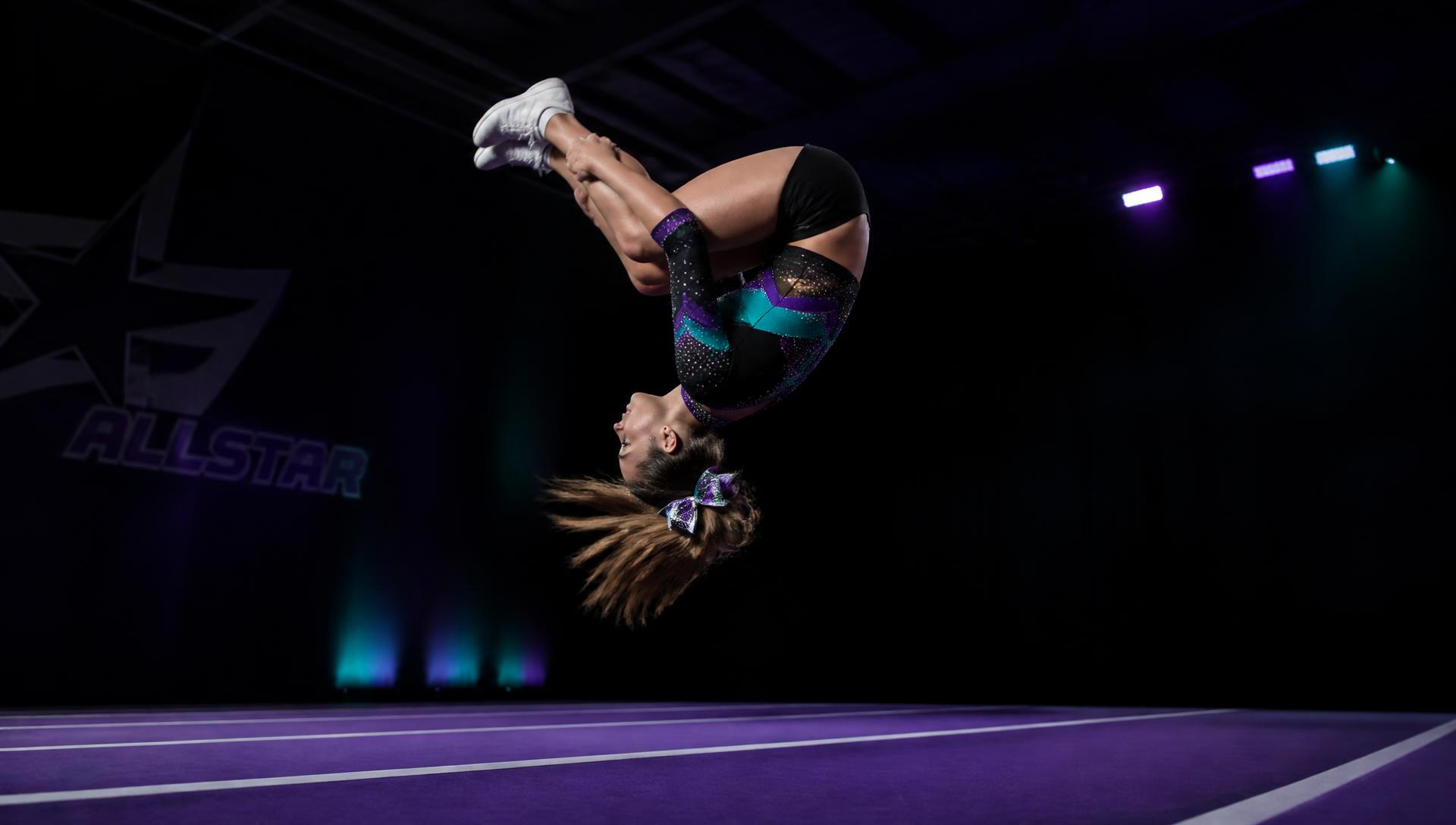 An Elite Athletics cheerleader at the apex of a back tuck above the gym's purple spring floor